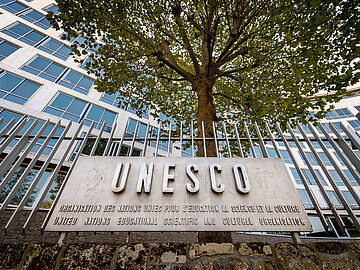 UNESCO Plakette Der Schriftzug UNESCO auf einer bronzenen Plakette, die an einem Stahlzaun befestigt ist. Im Hintergrund ein Baum und eine Gebäudefassade.