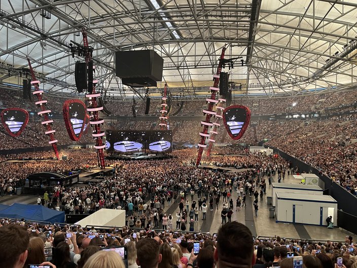 Bick von der Tribüne auf eine große Konzerthalle voll mit Menschen und einer Bühne in der Mitte.