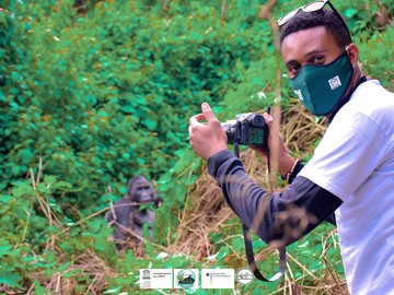 Ein Mann mit Mundschutz, der eine Kamera hält, blickt in die Kamera des Fotografen. Er möchte einen Gorilla fotografieren, der im Hintergrund des Bildes zwischen Sträuchern zu sehen ist. 