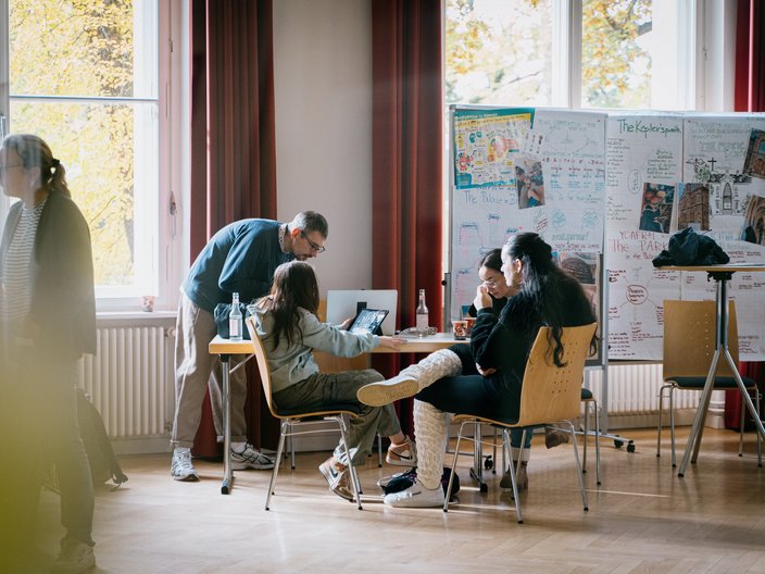 Eine Gruppe von Jugendlichen sitzt im Hintergrund an einem Gruppentisch und unterhalten sich. Dahinter stehen Pinnwände mit Plakaten.