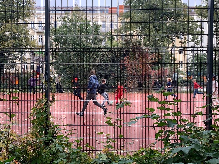 Perspektive auf einen Bolzplatz durch einen Gitterzaun. Im Hintergrund spielen Kinder unterschiedlichen Alters mit Ihren F&uuml;&szlig;en Ball.