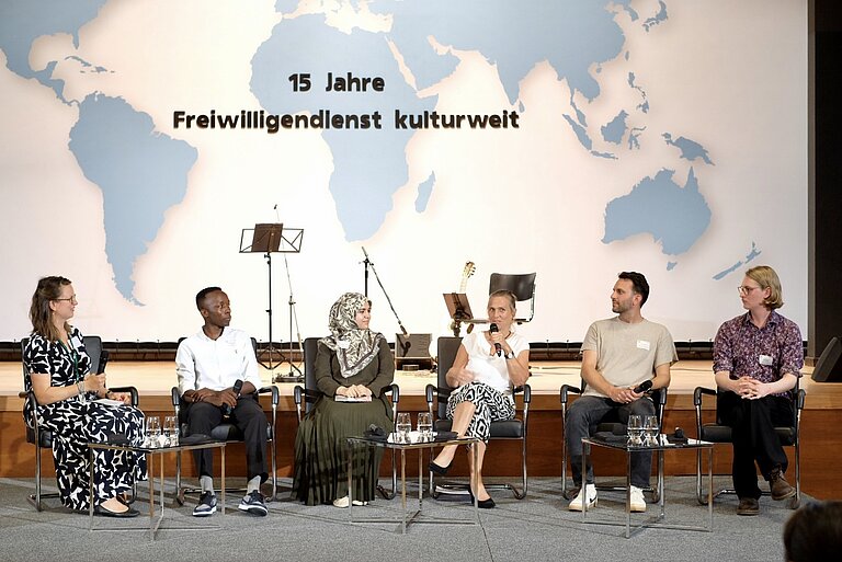 Sechs Personen auf einer B&uuml;hne sprechen miteinander, an der Wand dahinter ist 15 Jahre Freiwilligendienst kulturweit projeziert.