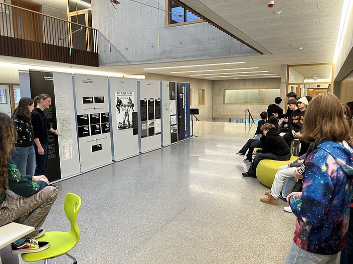 Schüler*innen stehen und sitzen um Pinnwände in der Schule herum welche Dokumente und Bilder im Bezug zum Holocaust präsentieren. 