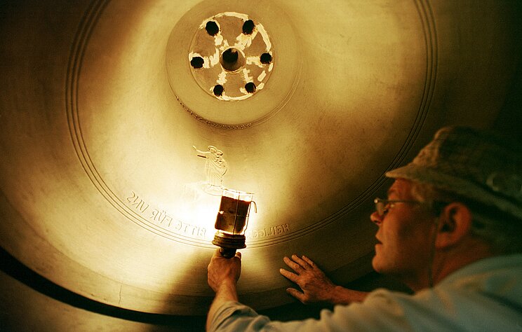 2_Glockenguss_Auflegen_Glockenzier Im Inneren einer großen Glocke legt ein Mann ein Glockenzier auf die Glocke auf.