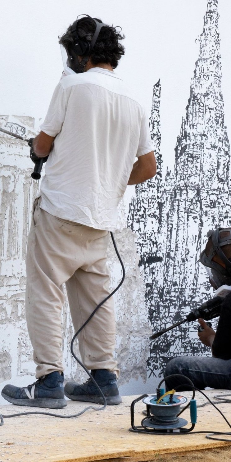 Zwei Künstler arbeiten an einem Wandbild. Sie fräsen mit Bohrmaschinen die Silhouetten von Gebäuden in eine Wand.