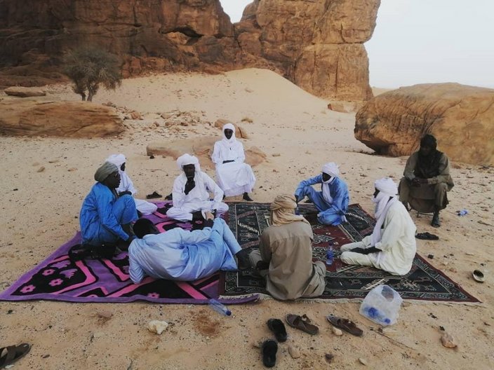 Touristenführer in traditioneller Kleidung sitzen auf rosa und lila Teppichen in der Wüste. Im Hintergrund sind die Felsen des Ennedi-Massivs. 