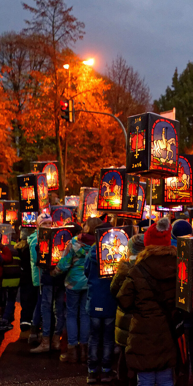 Kinder stehen auf der Stra&szlig;e im Halbdunklen und halten ihre Laternen in der Hand. Auf allen Laternen ist St. Martin auf einem Pferd abgebildet