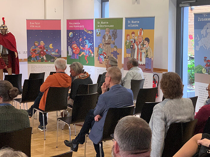 03_Martinsbräuche_ausstellung_und_vortrag_rene-bongart Person als St. Martin verkleidet steht in einem Konferenzsaal. Vor ihm sitzen Personen mit dem Rücken zur Kamera in Konferenzstühlen