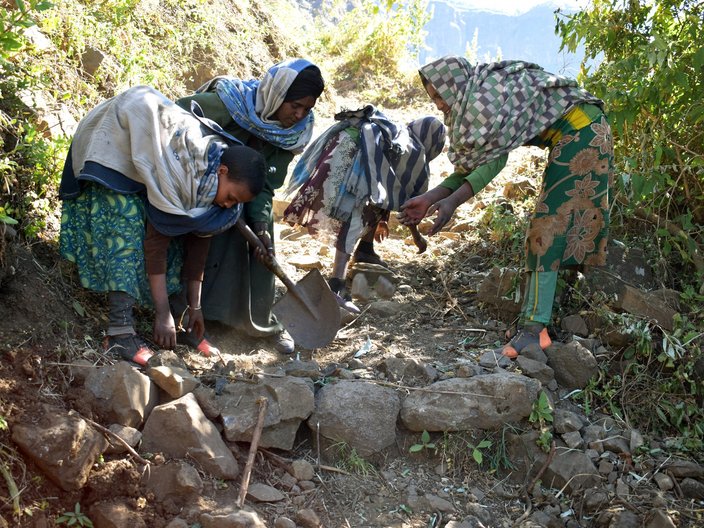 Vier Frauen bessern einen schmalen Wanderweg aus. Dazu benutzen sie Steine und Schaufeln. Sie tragen traditionelle Kleidung. 