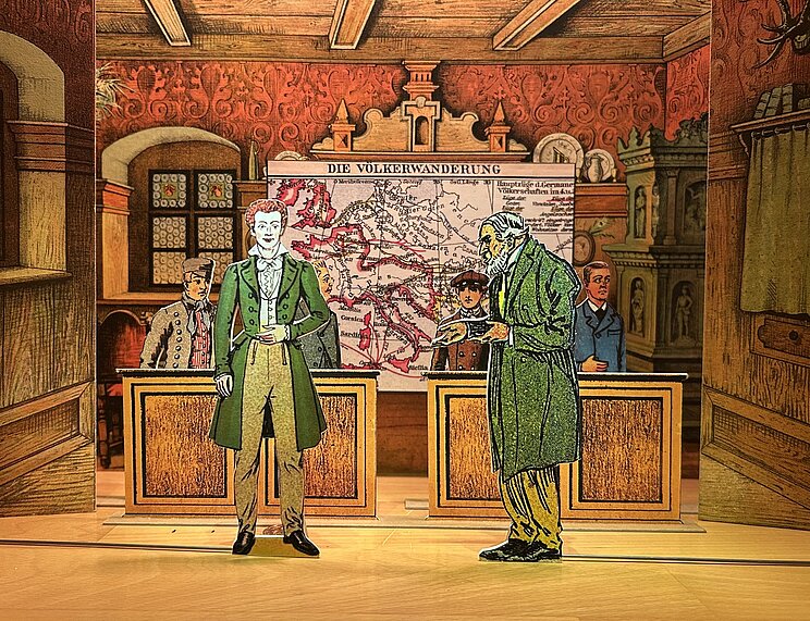 Papiertheater_Veranstaltung Historisch gestaltete Museums- oder Dioramaszene in einem holzverkleideten Raum. Im Vordergrund stehen zwei ausgeschnittene Figuren in historischer Kleidung vor Holztischen. Im Hintergrund befindet sich eine große Karte mit der Überschrift „Die Völkerwanderung“, auf der Wanderungsbewegungen in Europa eingezeichnet sind. Weitere Figuren sind im Raum verteilt. Die Einrichtung umfasst verzierte rote Tapeten, Holzbalken an der Decke, ein Fenster sowie einen kunstvoll gestalteten Kachelofen. Die Szene wirkt wie ein dreidimensionales Schaubild zur europäischen Geschichte.