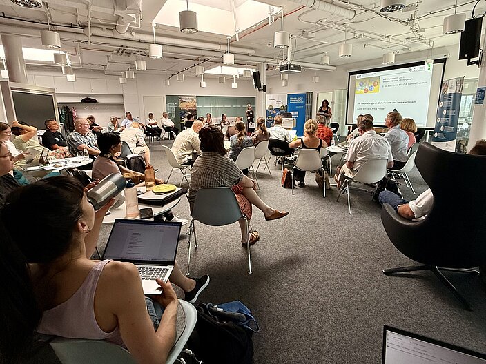 Panoramaperspektive auf Workshopraum mit Teilnehmenden, die auf Stühlen sitzen und Richtung einer Präsentation auf Leinwand schauen