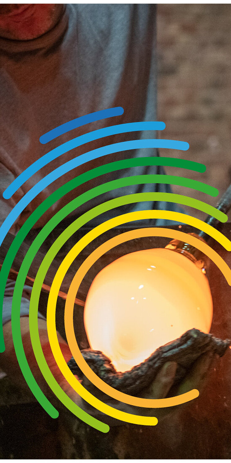 In einer Glasbl&auml;serei bearbeiten mehrere Personen mit langen Metallrohren eine gl&uuml;hende Glasmasse. Im Vordergrund leuchtet eine kugelf&ouml;rmige Glasblase in einem Ofen. &Uuml;ber dem Motiv liegt das farbige Logo zum Welttag des Immateriellen Kulturerbes. Rechts ist der Schriftzug &bdquo;Welttag des Immateriellen Kulturerbes&ldquo; zu sehen.