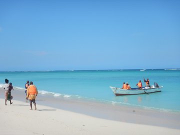 Blaues Meer, Sandstrand und Personen in einem Boot. Drei Personen laufen am Strand entlang.