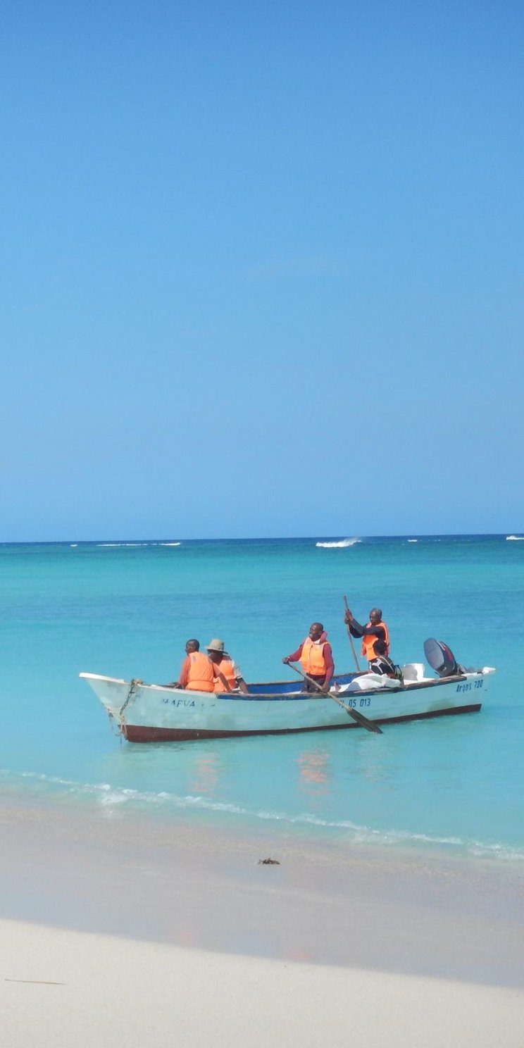 Blaues Meer, Sandstrand und Personen in einem Boot. Drei Personen laufen am Strand entlang.