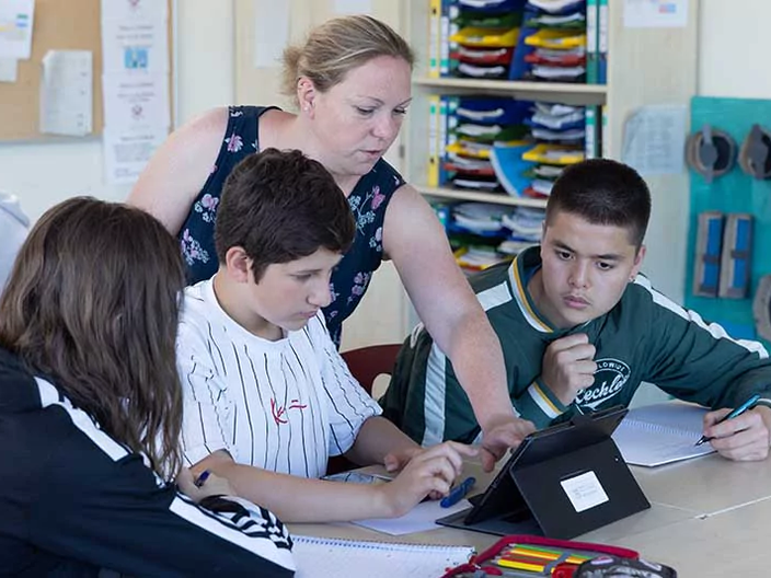 Drei Schüler sitzen an einem Tisch und die Lehrerin erklärt Ihnen etwas an einem Notebook.