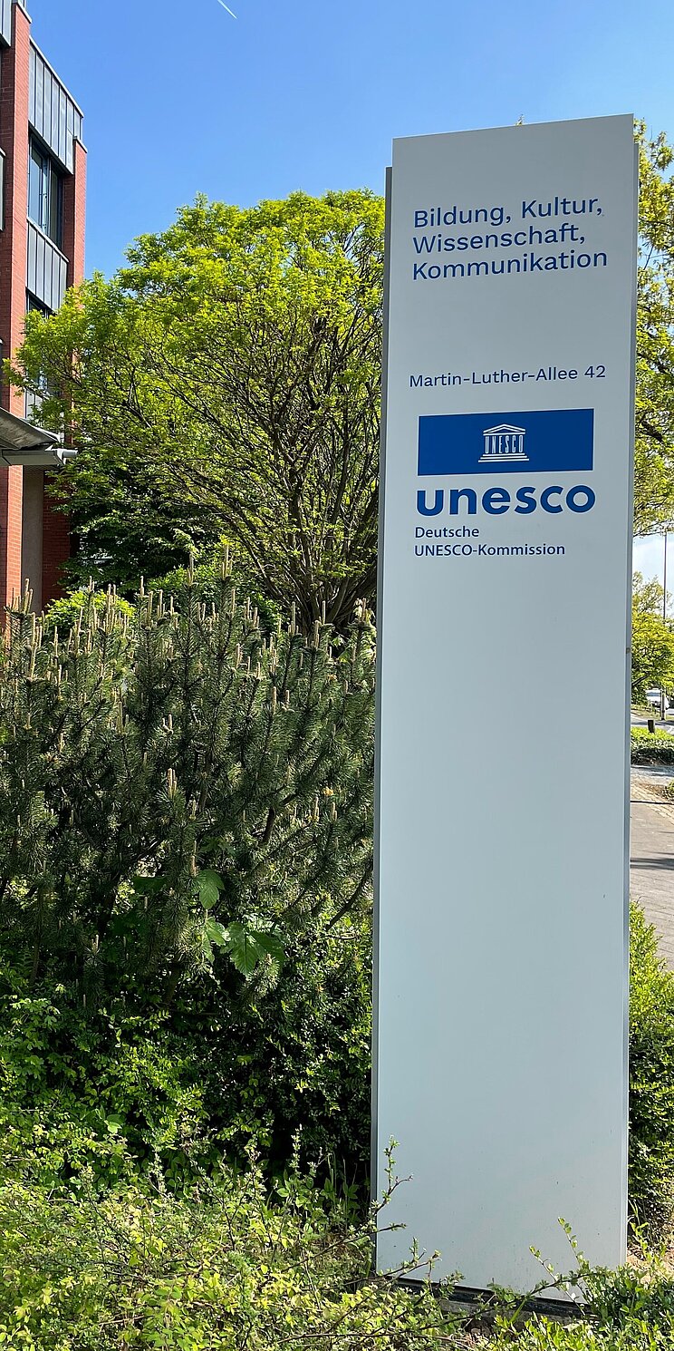 Wei&szlig;e Stele mit blauem UNESCO-Logo und Adresse vor dem DUK-Geb&auml;ude