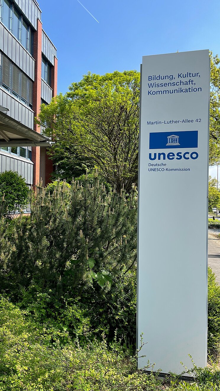 Wei&szlig;e Stele mit blauem UNESCO-Logo und Adresse vor dem DUK-Geb&auml;ude