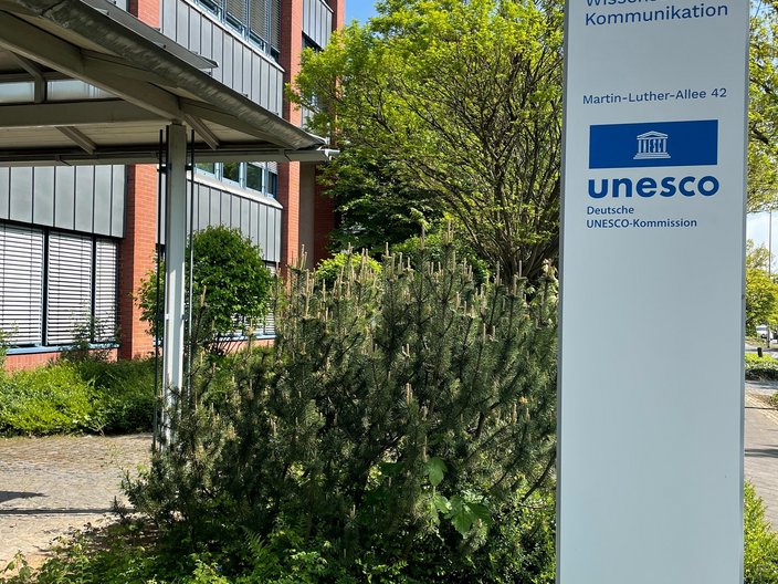 Weiße Stele mit blauem UNESCO-Logo und Adresse vor dem DUK-Gebäude