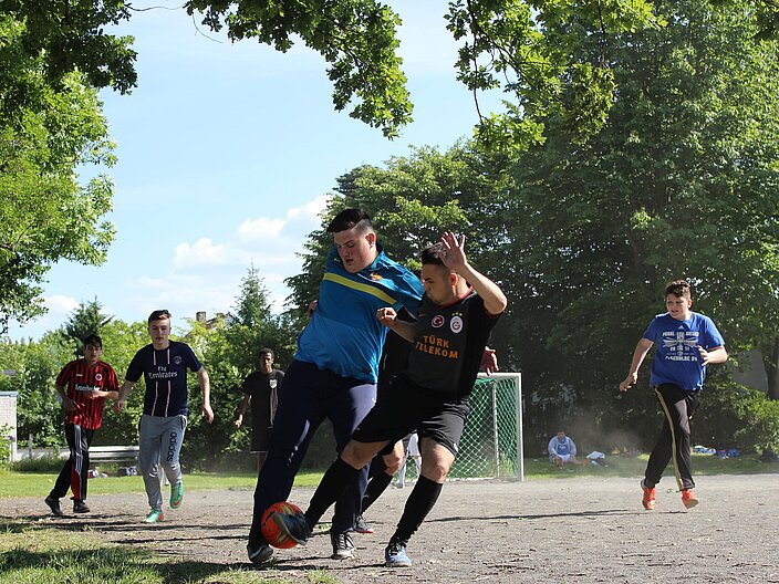 F&uuml;nf Jugendliche in Sportklamotten auf einem Schotterplatz. Im Vordergrund versuchen zwei Jugendliche mit ihren F&uuml;&szlig;en einen Fu&szlig;ball f&uuml;r sich zu gewinnen