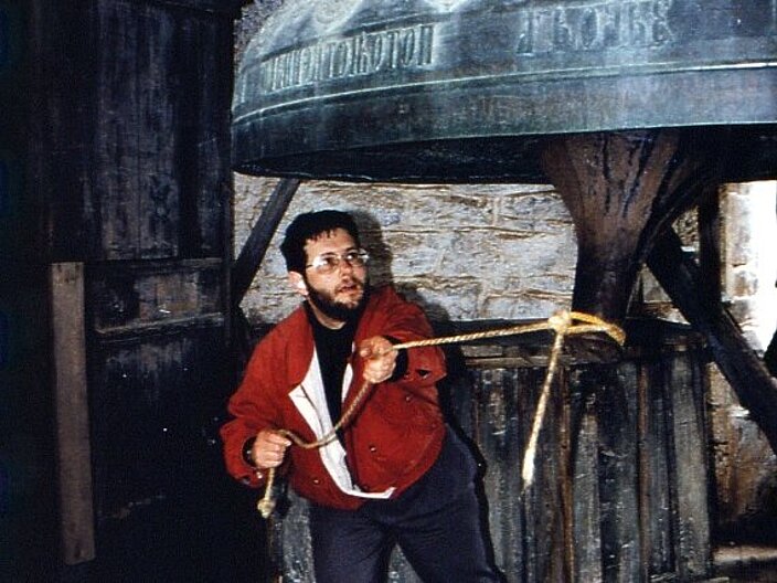 Person schlägt eine große Glocke in einem Glockturm mithilfe eines Seils an.