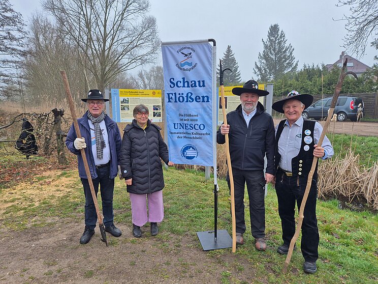 Flößerei1_Thiel Vier Personen stehen an einem Wassergraben neben einem Banner des Fördervereins Elsterfloßgraben mit der Aufschrift „Schau Flößen“. Auf dem Banner wird die Flößerei als Immaterielles Kulturerbe der Menschheit bezeichnet. Drei Personen halten lange Holzstangen. Im Hintergrund befinden sich Informationstafeln, Bäume und Sträucher.