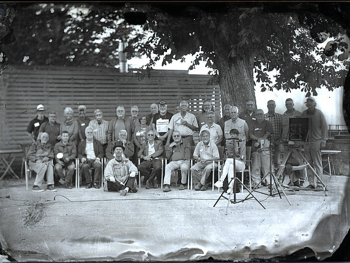 Schwarz-Weiß Foto von einer Menschengruppe, die für die Kamera posiert. Zum Teil sitzend auf Stühlen oder stehend dahinter.