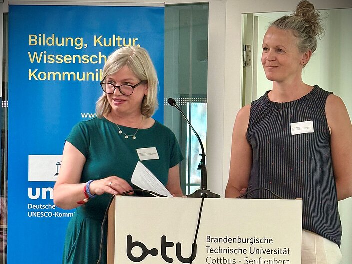 Katrin Seitz und Antje Reppe stehen gemeinsam am Redepult und hören einem Teilnehmenden zu.