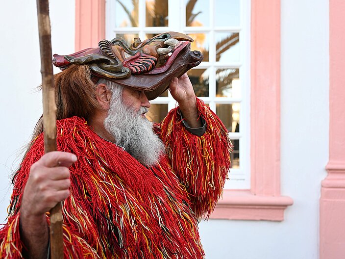 Eine Person mit Kostüm und Stock, die gerade ihre Holzmaske abnimmt, in Nahaufnahme von der Seite.