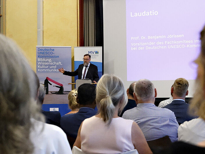 Vorsitzender des Fachkomitees Benjamin J&ouml;rissen h&auml;lt eine Laudatio auf der B&uuml;hne. Das Publikum schaut ihn an