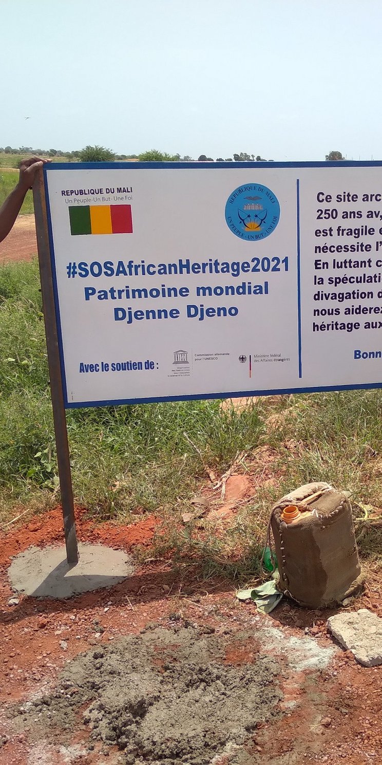 Drei Männer bauen eine Informationstafel auf, auf dem groß #SOSAfricanHeritage 2021 geschrieben ist. Hier wird auf Französisch das Projekt erklärt. 