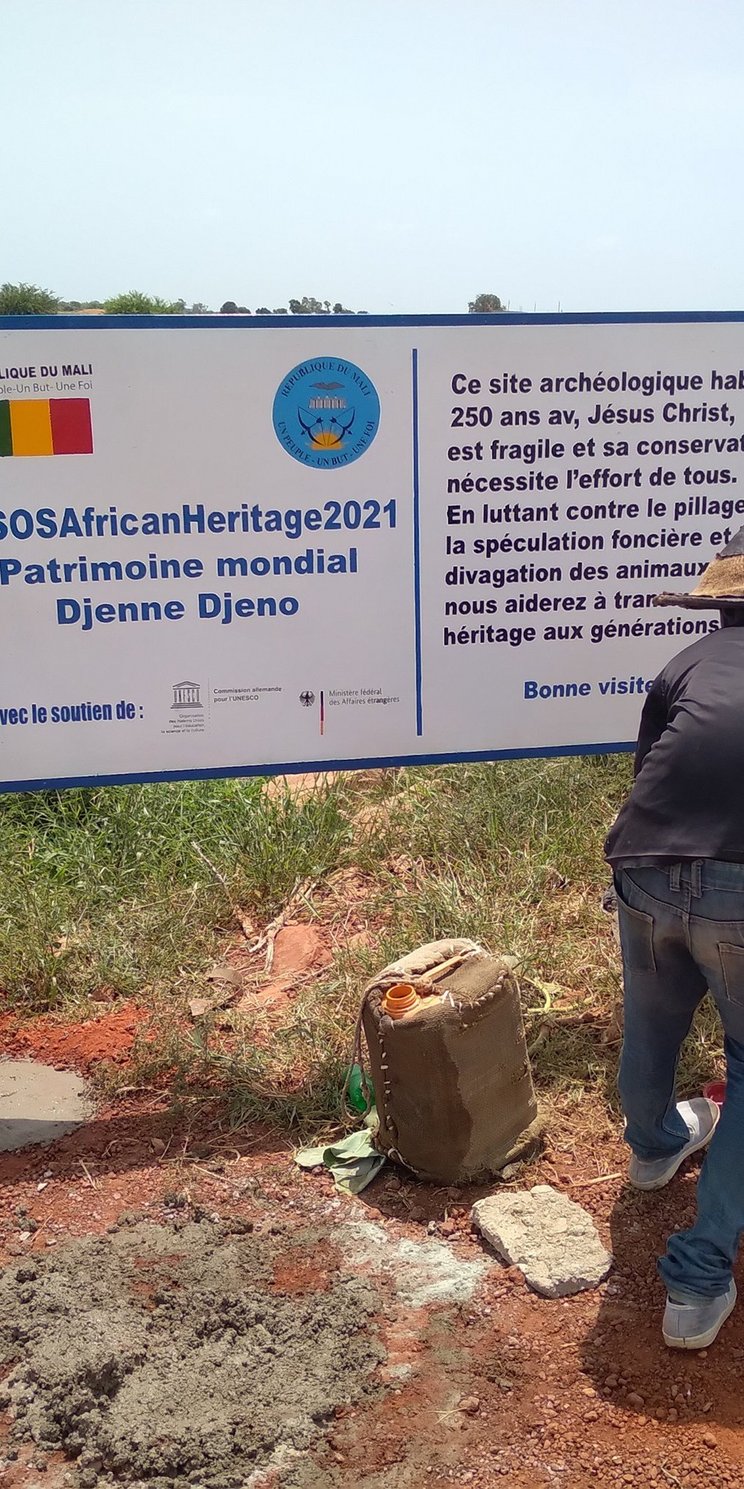 Drei Männer bauen eine Informationstafel auf, auf dem groß #SOSAfricanHeritage 2021 geschrieben ist. Hier wird auf Französisch das Projekt erklärt. 