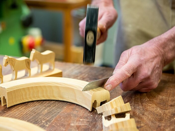 Mit einem Messer und Hammer werden aus einem Holzbogen Holzfiguren herausgebrochen.