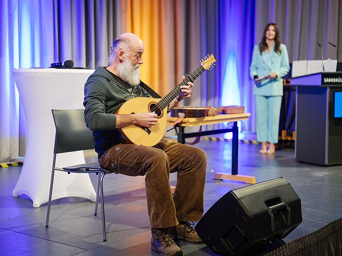 Urkundenverleihung_25_Waldzither Musiker sitzt auf Bühne mit Waldzither in der Hand und spielt ein Lied.