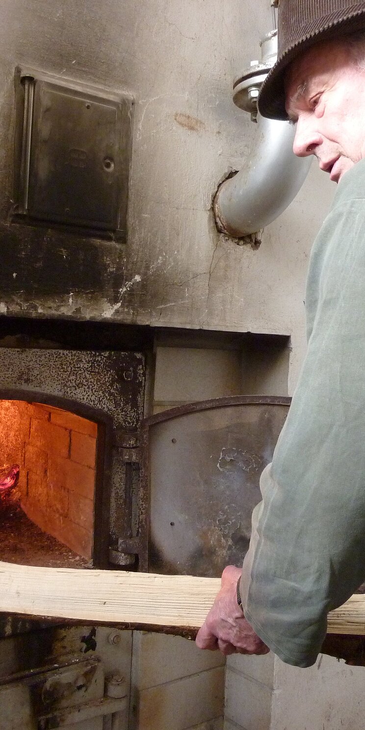 015-BY-Q-ZOIGLKULTUR-Foto001_TraditionelleBefeuerungKommunbrauhaus_Neuhaus Eine Person steht im Kommunbrauhaus in Neuhaus an einem offenen Befeuerungsofen und schiebt ein Holzscheit hinein.