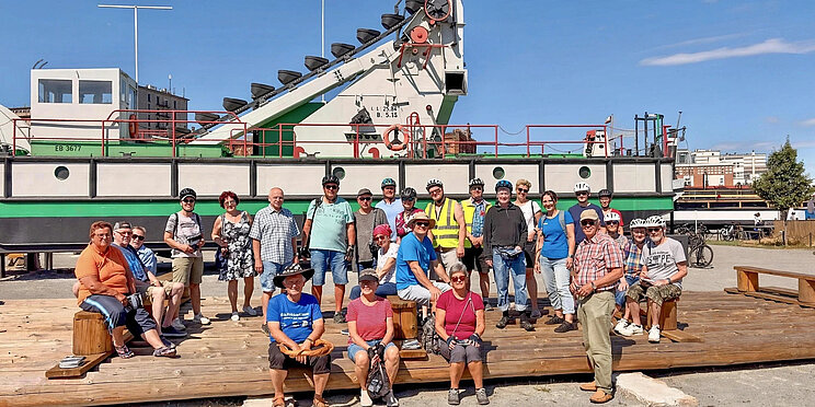 Eine gr&ouml;&szlig;ere Gruppe von Personen steht und sitzt auf einer Holzfl&auml;che vor einem wei&szlig;-gr&uuml;nen Arbeitsschiff. Einige Personen tragen Fahrradhelme und Fahrradkleidung. Im Hintergrund sind Geb&auml;ude und ein blauer Himmel zu sehen.