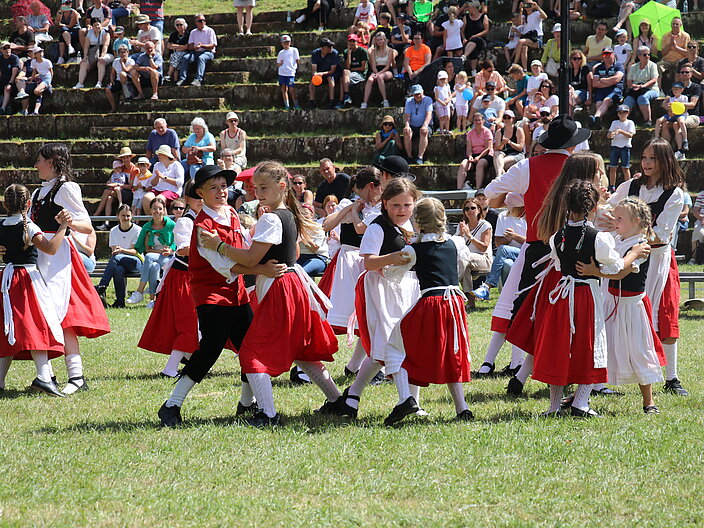 SCHÄFERLAUFFoto011_Kinder_beim_Tanz Mehrere Kinder tanzen paarweise auf einer Wiese. Sie tragen traditionelle Trachten. Im Hintergrund stehen Erwachsene und beobachten den Tanz.