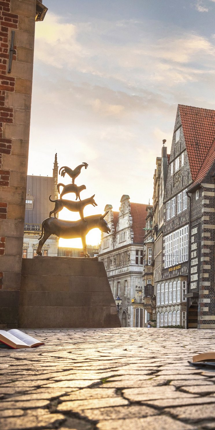 Die Bremer Stadtmusikanten neben dem Bremer Rathaus und aufgeschlagenen Büchern auf dem Kopfsteinpflaster