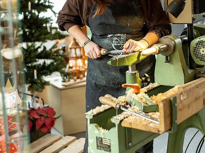 Eine Frau steht in der weihnachtlich geschmückten Schauwerkstatt und arbeitet an einem Holz-Werkstück.