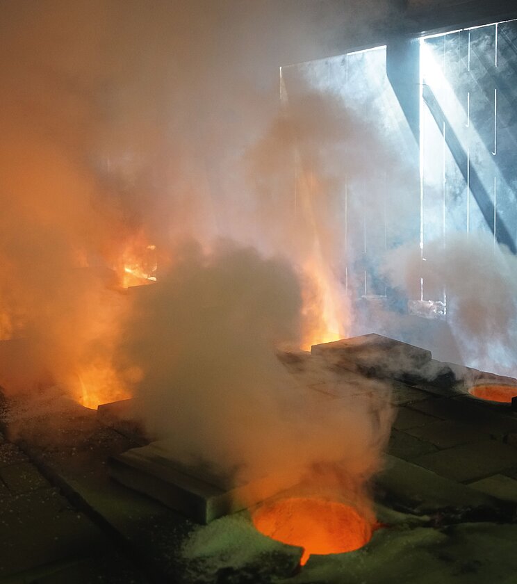 Kannofen_Veranstaltung Im Inneren einer Industrieanlage glüht geschmolzenes Metall in mehreren offenen Ofenöffnungen. Dichte weiße und graue Dampfschwaden steigen auf und füllen den Raum. Das orangefarbene Leuchten der Hitze kontrastiert mit kühlem Tageslicht, das durch große Öffnungen oder Fenster im Hintergrund einfällt. Dunkle Metallplatten und technische Anlagen umgeben die glühenden Öffnungen und vermitteln den Eindruck eines aktiven Schmelz- oder Gießprozesses in einer Gießerei.
