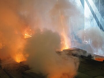 Im Inneren einer Industrieanlage gl&uuml;ht geschmolzenes Metall in mehreren offenen Ofen&ouml;ffnungen. Dichte wei&szlig;e und graue Dampfschwaden steigen auf und f&uuml;llen den Raum. Das orangefarbene Leuchten der Hitze kontrastiert mit k&uuml;hlem Tageslicht, das durch gro&szlig;e &Ouml;ffnungen oder Fenster im Hintergrund einf&auml;llt. Dunkle Metallplatten und technische Anlagen umgeben die gl&uuml;henden &Ouml;ffnungen und vermitteln den Eindruck eines aktiven Schmelz- oder Gie&szlig;prozesses in einer Gie&szlig;erei.