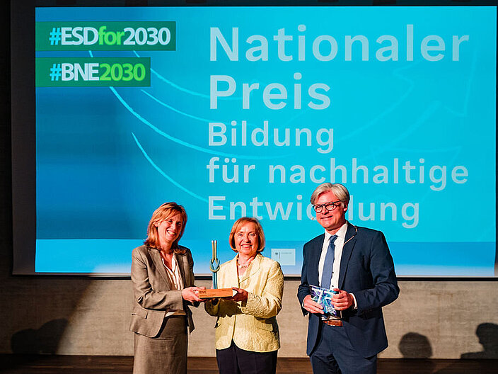 Karin Prien, Maria B&ouml;hmer, Roman Luckscheiter stehen auf der B&uuml;hne und halten einen Preis hoch.