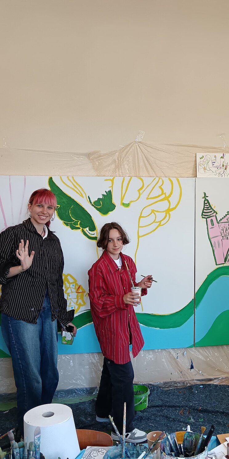 Vier ukrainische Jugendliche stehen zusammen mit einem Künstler vor einer großen zu bemalenden Leinwand in der Schule. Alle haben Pinsel in der Hand und auf der Leinwand entsteht bereits ein Kunstwerk in bunten Farben.