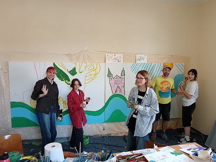 Vier ukrainische Jugendliche stehen zusammen mit einem Künstler vor einer großen zu bemalenden Leinwand in der Schule. Alle haben Pinsel in der Hand und auf der Leinwand entsteht bereits ein Kunstwerk in bunten Farben.