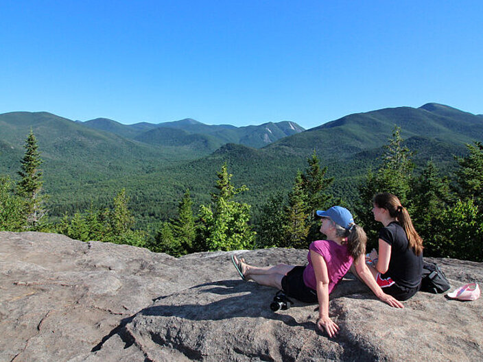 The picture shows two women sitting on a rock. Their backs are turned to the viewer and you are looking with them into the nature park where you can see the mountains in the background.