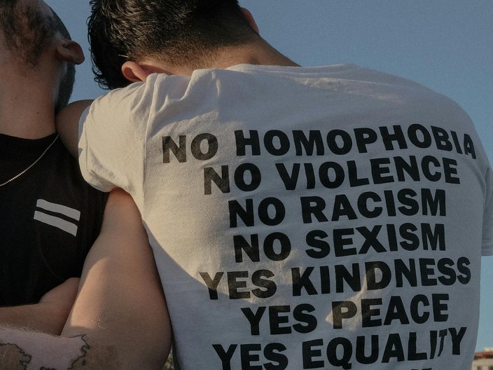 Zwei Männer lehnen aneinander. Auf dem T-Shirt-Rücken des einen Mannes steht "No homophobia, no violence, no racism, no sexism, yes kindness, yes peace, yes equality, yes love", 