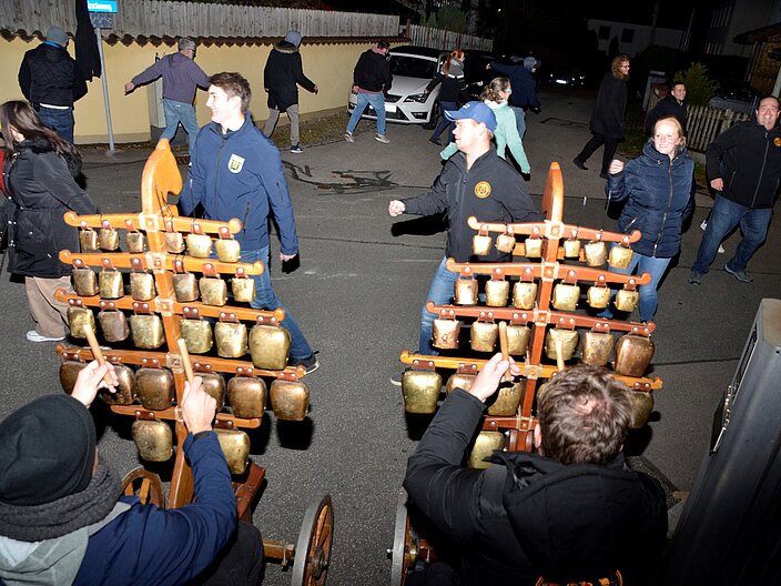 Die Mitwirkenden üben einen Kreistanz bei der Tanzprobe vor dem Perchtenlauf. Zwei Personen im Vordergrund spielen ein Glockenspiel.