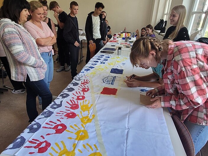 Ukrainische Schüler*innen und Lehrkräfte stehen in einem Klassenraum vor einem langen Tisch auf dem ein Leinentuch ausgelegt ist welches von Schüler*innen mit Handabtrücken und dem UNESCO-Zeichen bemalt bzw. gestempelt wird.