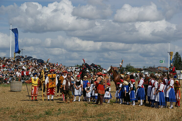 Gro&szlig;e Gruppe von Menschen in historischer Festkleidung auf einem offenen Feld. Vor Publikum pr&auml;sentieren sich Teilnehmende einer traditionellen Veranstaltung mit farbenfrohen Trachten, historischen Darstellungen und Reitenden unter bew&ouml;lktem Himmel.