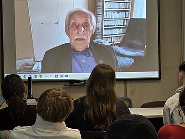In einem Klassenraum schaut eine Gruppe von Schülern und Schülerinnen auf die ausgefahrene Leinwand an der Wand über die Leon Weintraub aus Schweden zugeschaltet ist.