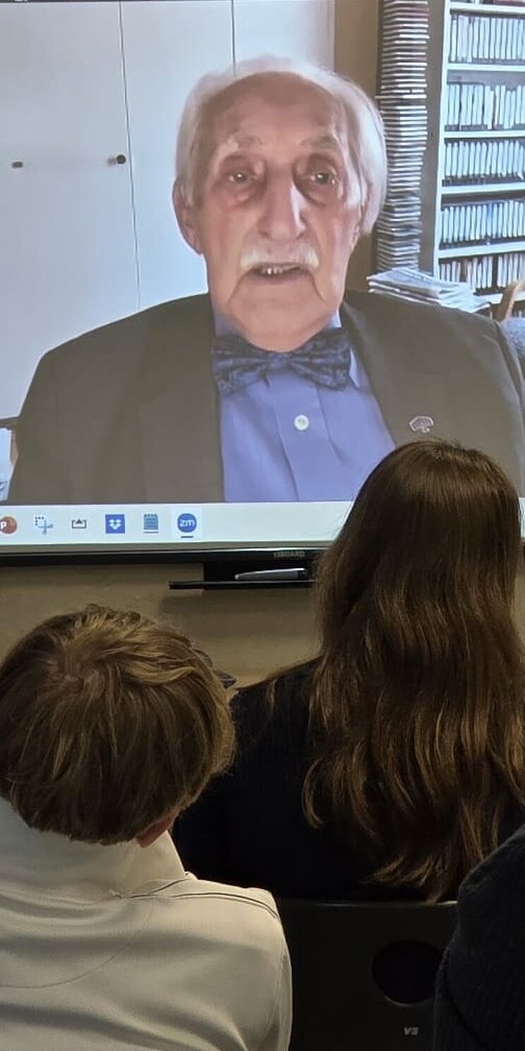 In einem Klassenraum schaut eine Gruppe von Schülern und Schülerinnen auf die ausgefahrene Leinwand an der Wand über die Leon Weintraub aus Schweden zugeschaltet ist.